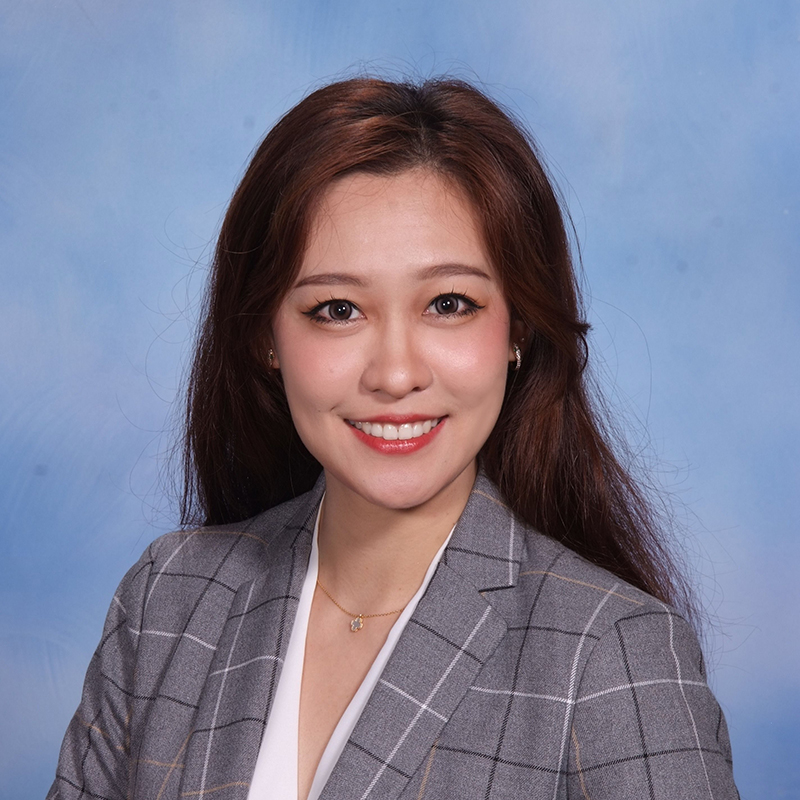 The image shows a woman posing for a portrait with a smile. She has dark hair, wears makeup, and is dressed in a professional attire consisting of a blazer over a shirt and a necklace.